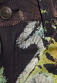 Tecido preto com uma estampa abstrata colorida apresentando padrões em verde, amarelo e branco. Inclui um botão de metal e detalhes de costura visíveis.