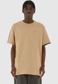 Beige bomulds t-shirt med løs pasform, korte ærmer og rund hals. Har lille rødt logo tekst på venstre brystområde.