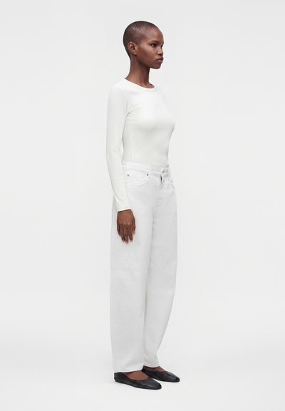 Femme aux cheveux courts portant un haut blanc à manches longues, un pantalon blanc large et des chaussures plates noires, debout sur un fond clair uni.