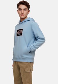 Svetlo modra kapuca iz mehkega blaga, z prednjim žepom in črnim logotipom.  Ujemajoči bež cargo hlače z bočnimi žepi.