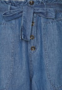 Denim overall med dragsko i midjan, knäppning fram och två stora sidofickor. Tyget är en enhetlig mörkblå med en mjuk textur.