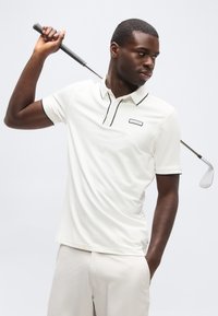 Homme en polo blanc et pantalon tenant un club de golf derrière son cou, regardant sur le côté sur un fond uni.