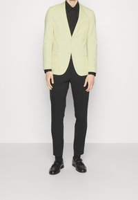 Zielona marynarka w kolorze limonki o dopasowanym kroju, z zapięciem na jeden guzik, klapami w typie notch, zestawiona z czarnymi spodniami slim fit i czarnymi butami.