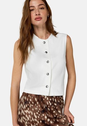 Kvinde med langt brunt hår iført hvid ærmeløs vest med knapper og brun nederdel med hvidt prikmønster, stående foran en ensfarvet baggrund.