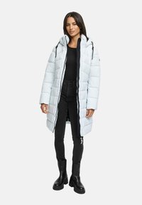 Mujer vestida con un abrigo acolchado largo azul claro con capucha, camiseta negra, jeans skinny negros y botas gruesas negras, de pie contra un fondo blanco.