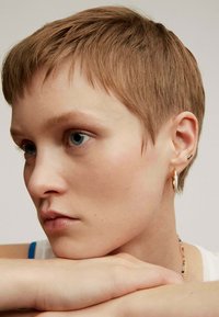 Pelo corto, color marrón claro; lleva un pendiente de aro dorado y un pequeño arete de perno. Tono de piel natural, ligeras pecas, manos descansando sobre los brazos.