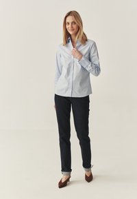 Camicia azzurro chiaro con maniche lunghe, abbinata a jeans di denim scuri, con polsini ripiegati e décolleté marroni a punta, presentata su uno sfondo neutro.