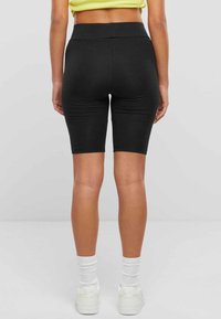 Schwarze Radshorts mit hohem Bund, die eng an den Oberschenkeln anliegen. Glatte Textur, keine sichtbaren Muster, kombiniert mit weißen Turnschuhen und Socken.