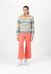 Fleece pullover met een meerkleurig golfpatroon, voorzien van een tan kraag en een front pocket. Gecombineerd met brede koraalkleurige corduroy broeken.
