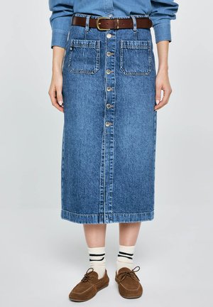 Dżinsowa spódnica midi z guziczkami z przodu i naszywanymi kieszeniami, noszona z brązowym paskiem, białymi skarpetkami z czarnymi paskami oraz brązowymi sznurowanymi butami.