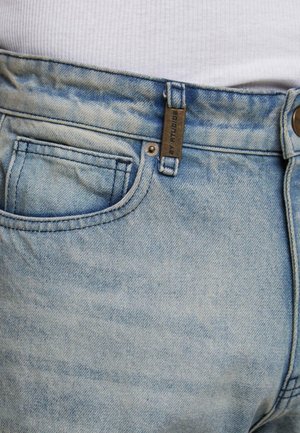 Šviesiai mėlynos džinsinės kelnės su juosmeniu ir priekiniais kišenėmis, kuriose yra metalinė etiketė, dėvimos su baltu koralu, matomu virš juosmens.