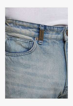 Šviesiai mėlynos džinsinės kelnės su juosmeniu ir priekiniais kišenėmis, kuriose yra metalinė etiketė, dėvimos su baltu koralu, matomu virš juosmens.