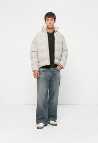 Lysegrå pufferjakke med høj krave, parret med en mørk strikket sweater og løse blå jeans. Fodtøjet har grå og hvide accenter.