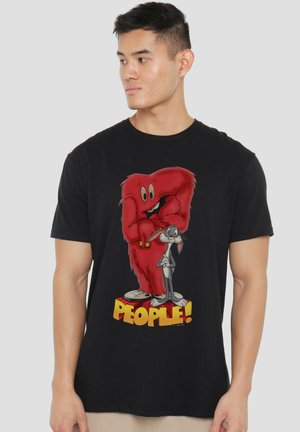 Junger Mann, der ein schwarzes T-Shirt mit den Cartoon-Figuren Gossamer und Bugs Bunny sowie dem Wort "PEOPLE!" in fetten gelben Buchstaben trägt.