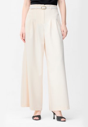Pantalon crème à jambes larges avec plis devant et ceinture assortie, porté avec des mules à talons ouvertes noires et un top noir.