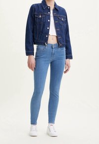 Chaqueta de mezclilla azul oscuro con diseño corto, combinada con jeans ajustados azul claro y zapatillas blancas. Textura suave, detalles de botones visibles.