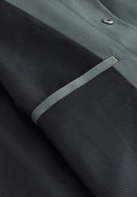 Gros plan d'une poche en tissu noir avec un motif texturé, bordée de tissu gris et un bouton gris au-dessus de la poche.