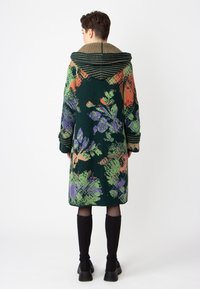 Long green knit coat featuring a multicolored floral pattern. Has a striped brown hood and wide sleeves. Accented with ribbed cuffs and hem.