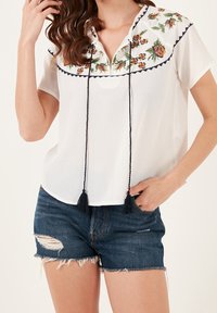 Blouse blanche avec broderies florales et accents bleu marine, manches courtes, et cordon à pampilles. Associée à un short en denim déchiré.