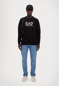 Juodas džemperis su logotipu, mėlyni džinsai, balti sportbačiai ir juodas kepurė. Mažas juodas sagūnos krepšys nešiojamas ant dešinės peties.