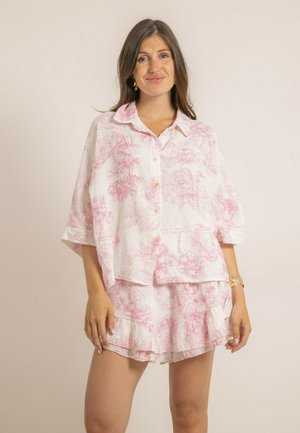 Femme portant une chemise blanche ample à boutons et un short assorti avec des motifs floraux roses, debout devant un fond beige uni.