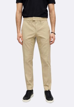 Uomo che indossa pantaloni chinos beige, maglietta nera, braccialetto nero e sneaker nere con suola bianca, in piedi con le braccia rilassate lungo i fianchi.