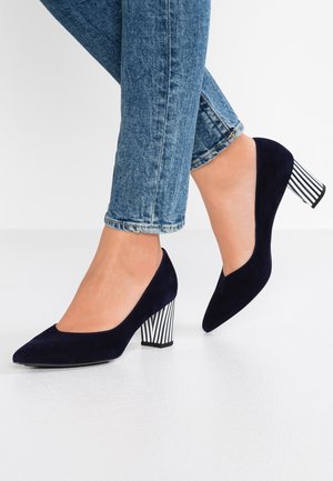 Kvinde iført mørkeblå ruskindspumps med tykke sort-hvide stribede hæle og blå skinny jeans.