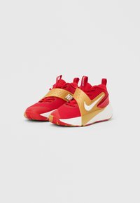 Τα αθλητικά παπούτσια Nike διαθέτουν κόκκινο επάνω μέρος από πλέγμα με λουράκι σε χρυσό χρώμα, λευκή ενδιάμεση σόλα και υφασμένο κάτω μέρος σόλας. Ο σχεδιασμός περιλαμβάνει ένα εμφανές λογότυπο.