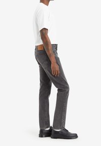 Jeans de mezclilla gris con un corte recto, que presentan un parche trasero de cuero marrón y un desvanecimiento sutil, combinados con zapatos negros y una camiseta blanca.