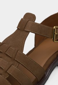 Καφέ σουέντ σανδάλι με πολλαπλές λουρίδες, κλείσιμο με τοκά στην κορυφή και ραμμένες λεπτομέρειες πάνω σε ανοιχτό φόντο.