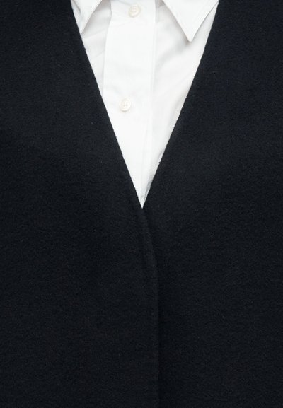 Μαύρο γιλέκο από μάλλινο ύφασμα με βαθύ V-λαιμόκοψη, διαθέτει απαλό υφή και ελάχιστα μεταλλικά στοιχεία, φορεμένο πάνω από ένα λευκό πουκάμισο με γιακά.