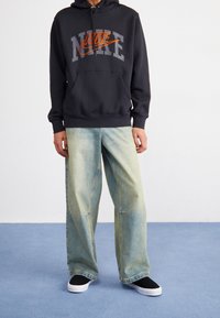 Svart hoodie med grå och orange "NIKE"-logotyp, kombinerad med ljusblå baggy jeans och svarta slip-on skor. Strukturerad tyg, avslappnad passform.