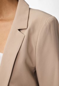 Béžový blazer se strukturovaným klopem a hladkým textilním povrchem. Detailní záběr zdůrazňující splývání látky a jemný lesk.