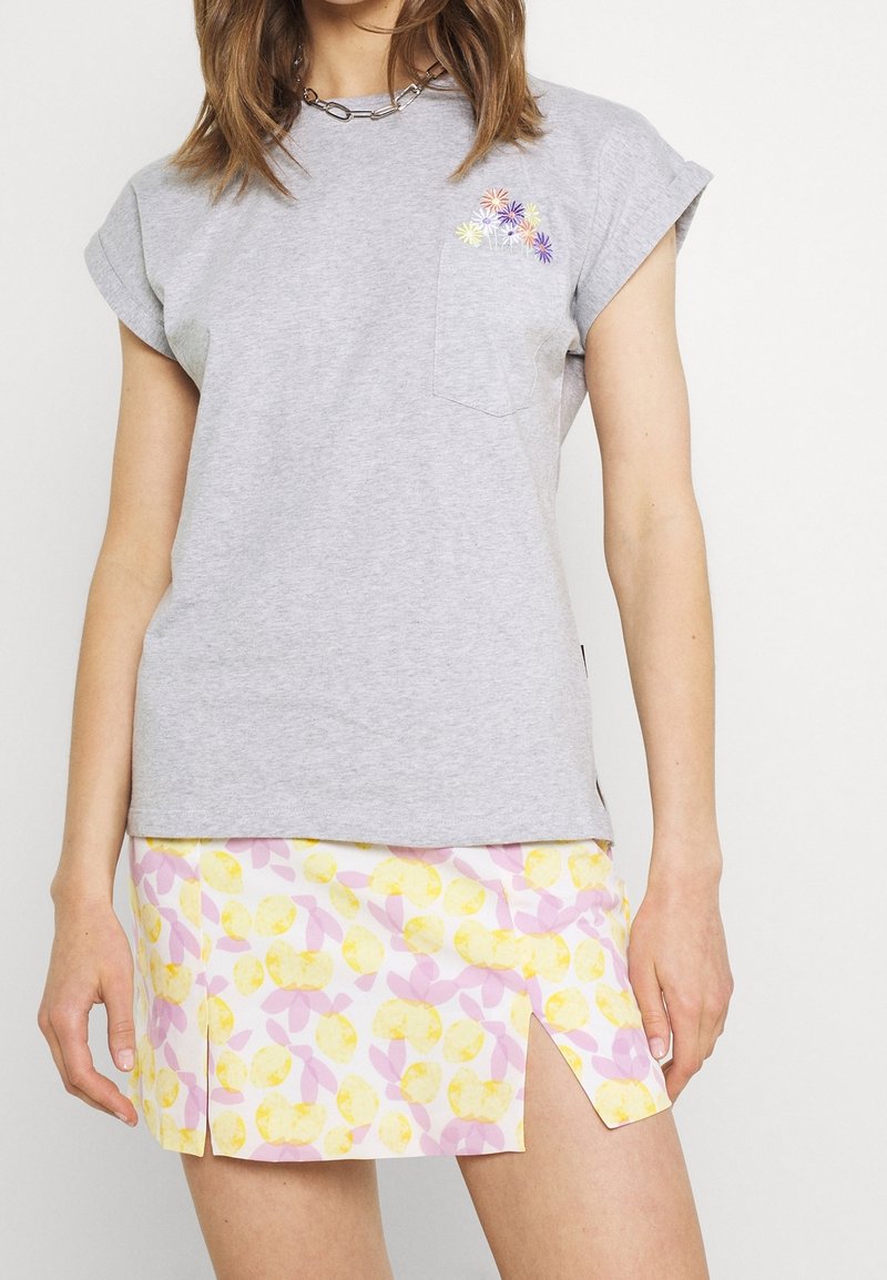 Femme portant un t-shirt gris avec broderie florale sur la poche et une jupe blanche à motif floral jaune et rose avec des fentes à l'avant.