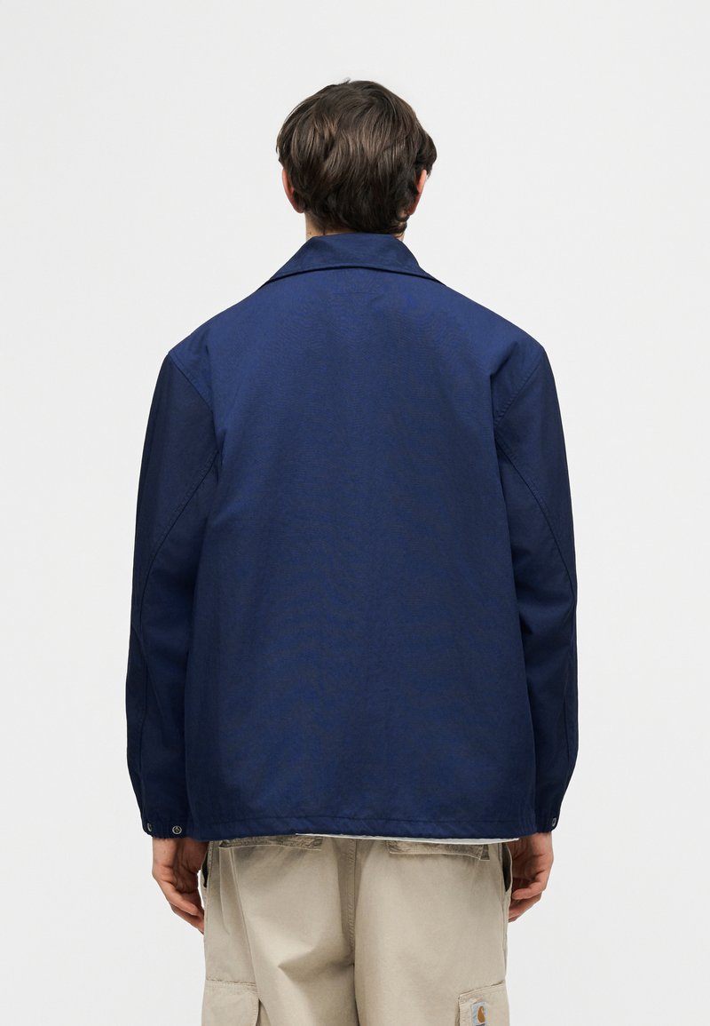 Uomo con giacca blu navy e pantaloni cargo beige, visto di spalle su uno sfondo semplice grigio chiaro.