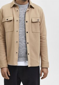 Man med beige knappad jacka över en grå stickad tröja och vit skjorta, med svarta byxor mot en plain ljus bakgrund.