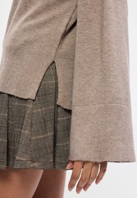 Μπεζ πλεκτό πουλόβερ με φαρδιές μαν�ίκες και πλευρικές σχισμές, συνδυασμένο με φούστα πλισέ σε καρώ σχέδιο σε σκούρες αποχρώσεις και φωτεινές λεπτομέρειες.