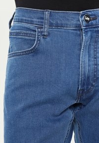 Ljust blå denimjeans har en klassisk femfickdesign, en knappstängning och kontrasterande sömmar längs sömmarna.