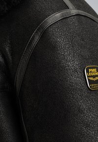 Close-up van de schouder van een zwart, textured leren jack met een gestikt leren afwerking en een gele PME Legend merkpatch.