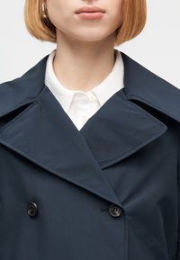 Γυναίκα με σκούρο μπλε διπλόμπουφο παλτό πάνω σε λευκό πουκάμισο με γιακά, εμφανίζοντας τα χείλη και το κάτω μέρος του προσώπου της με κοντά ξανθά μαλλιά.