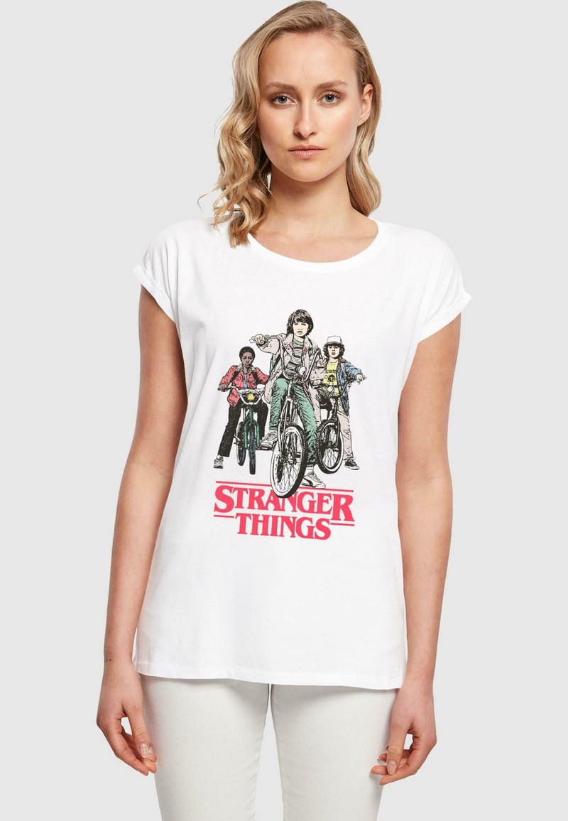 Valge puuvillane t-särk, millel on graafiline print kolmest tegelasest jalgrattatel, allpool illustratsiooni on kirjas "STRANGER THINGS" punases kirjas.