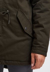 Olivgrön parka-jacka med dragsko stängning, knappförsedda ficklock och slät tygtextur med synliga sömnadsdetaljer.