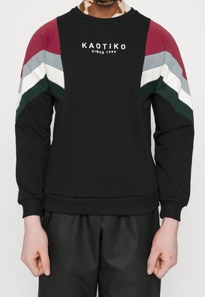 Person trägt schwarzen Sweatshirt mit roten, grauen, weißen und grünen Chevron-Streifen an den Ärmeln und dem Schriftzug „KAOTIKO SINCE 1999“ auf der Brust.