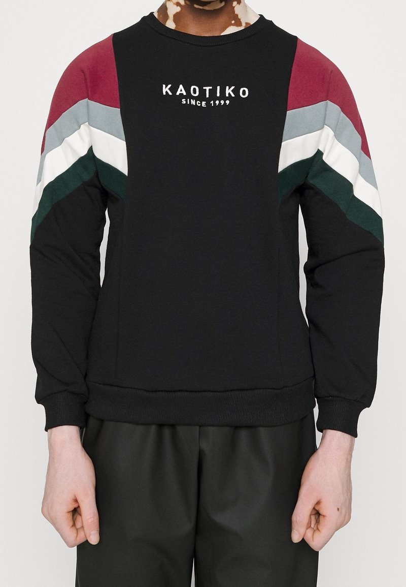 Personne portant un sweat-shirt noir avec des bandes chevrons rouges, grises, blanches et vertes sur les manches et le texte « KAOTIKO SINCE 1999 » sur la poitrine.