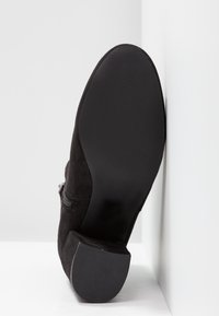 Botín negro de ante hasta el tobillo con suela de goma texturizada, detalle de cremallera lateral y tacón afilado. La base tiene un acabado suave con ligeras estrías.