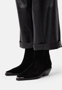 Botine din suede negru cu vârful ascuțit și toc conic. Prezentând un accent metalic pe capacul vârfului. Asortate cu pantaloni din piele neagră.