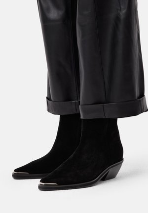 Stivaletti neri in suede con punta affusolata e tacco stretto. Presentano un accento metallico sulla punta. Abbinati a pantaloni in pelle neri.