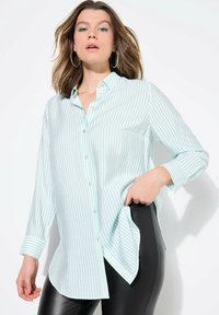 Camisa de botones de manga larga a rayas azul claro y blanca, con un bolsillo, dobladillo curvado y un corte relajado, combinada con pantalones negros.