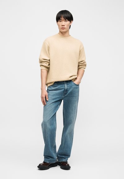 Jeune personne aux cheveux courts noirs portant un pull beige, un jean bleu ample et des chaussures marron foncé, debout devant un fond uni.