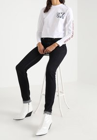 Weißes Langarmshirt mit "CK"-Logo, kombiniert mit schwarzen Jeans und weißen Lackleder-Ankle-Boots mit Wedge-Absätzen, auf einem weißen Hocker.